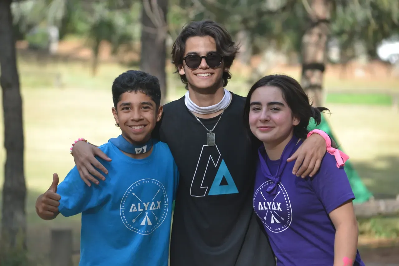 Tres jóvenes sonrientes con playeras de ALYAX abrazados en el campamento